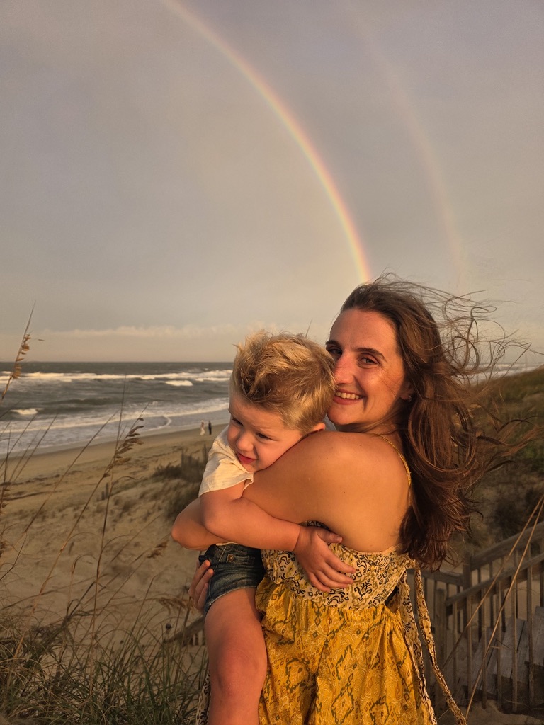 Rachel holding a toddler on the beach beneath a rainbow
